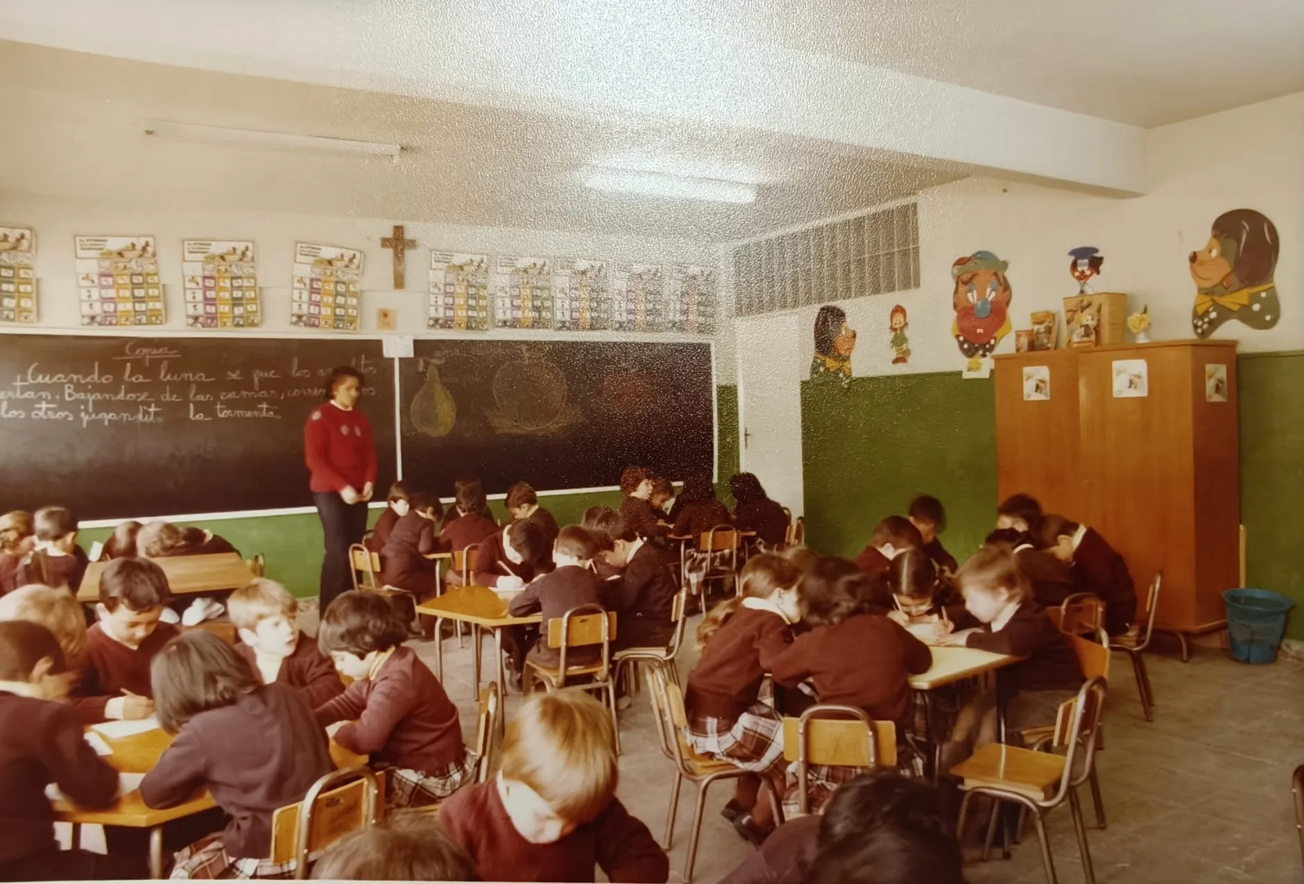 Una clase trabajando en sus mesas, con la pizarra escrita y un crucifijo en la pared